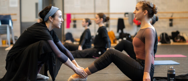 In a large, industrial-looking dance studio, Isabella McGuire Mayes squats down and holds the ankles of an adult ballet student as she sits up tall, bends her knees up and poins her toes. They both wear dance clothing and are surrounded by other dancers doing the same exercise.