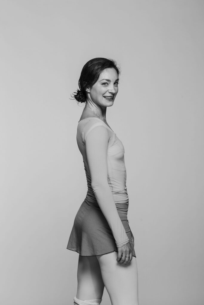 (In black and white) Amy Watson stands in profile in ballet attire, her dark hair in a low bun, and smiles toward the camera with her arms relaxed by her side.