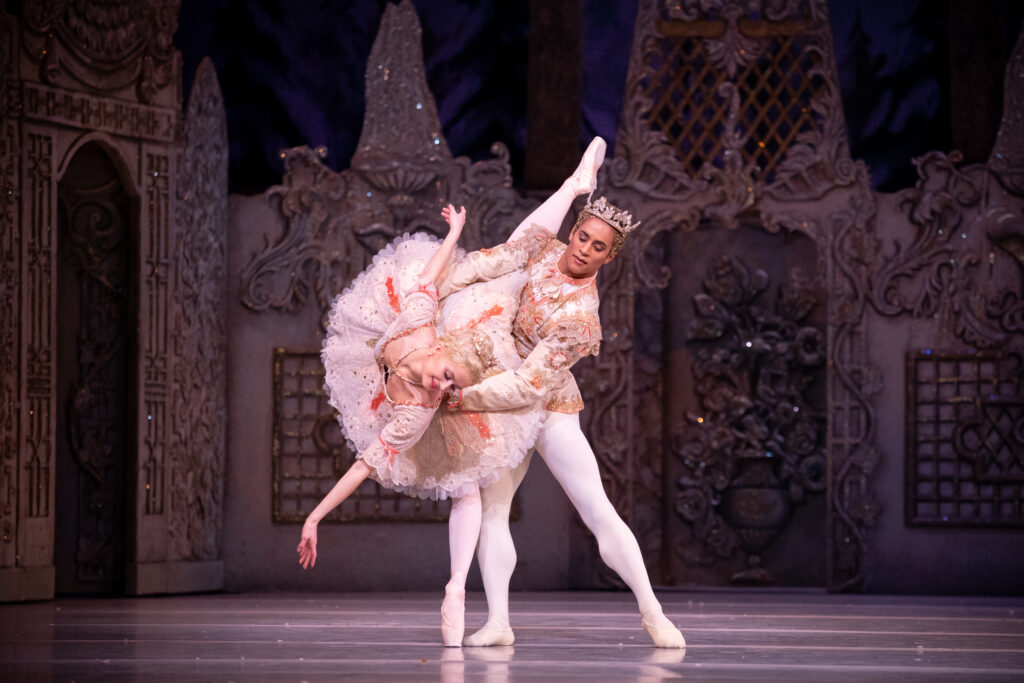 Anna Rose O'Sullivan, wearing an intricatly detailed pink tutu, does a penché in arabesque as Marcelina Sambé holds her by the waist with his left leg in tendu. She sweeps her left arm across and twists her upper body slightly, looking at her hand. Sambé wears a matching detailed tunic, white tights and slippers, and a large crown.