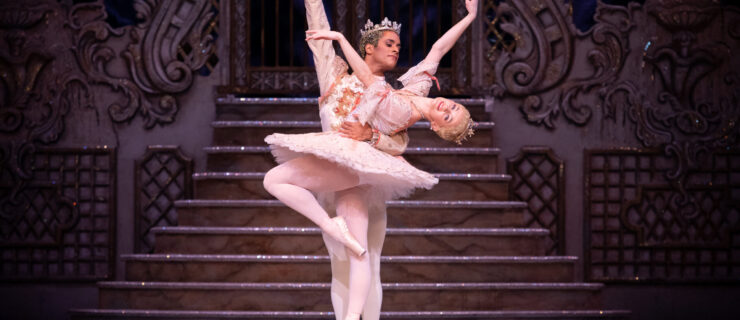 Anna Rose O'Sullivan poses in parralel passee on pointe, bending her upper body back in cambé with her arms in third arabesque. Marcelino Sambé stands beihnd her and holds her by the waist. She wears a pink tutu and gold tiara, while he wears a pink tunic, white tights and slippers, and a large crown.