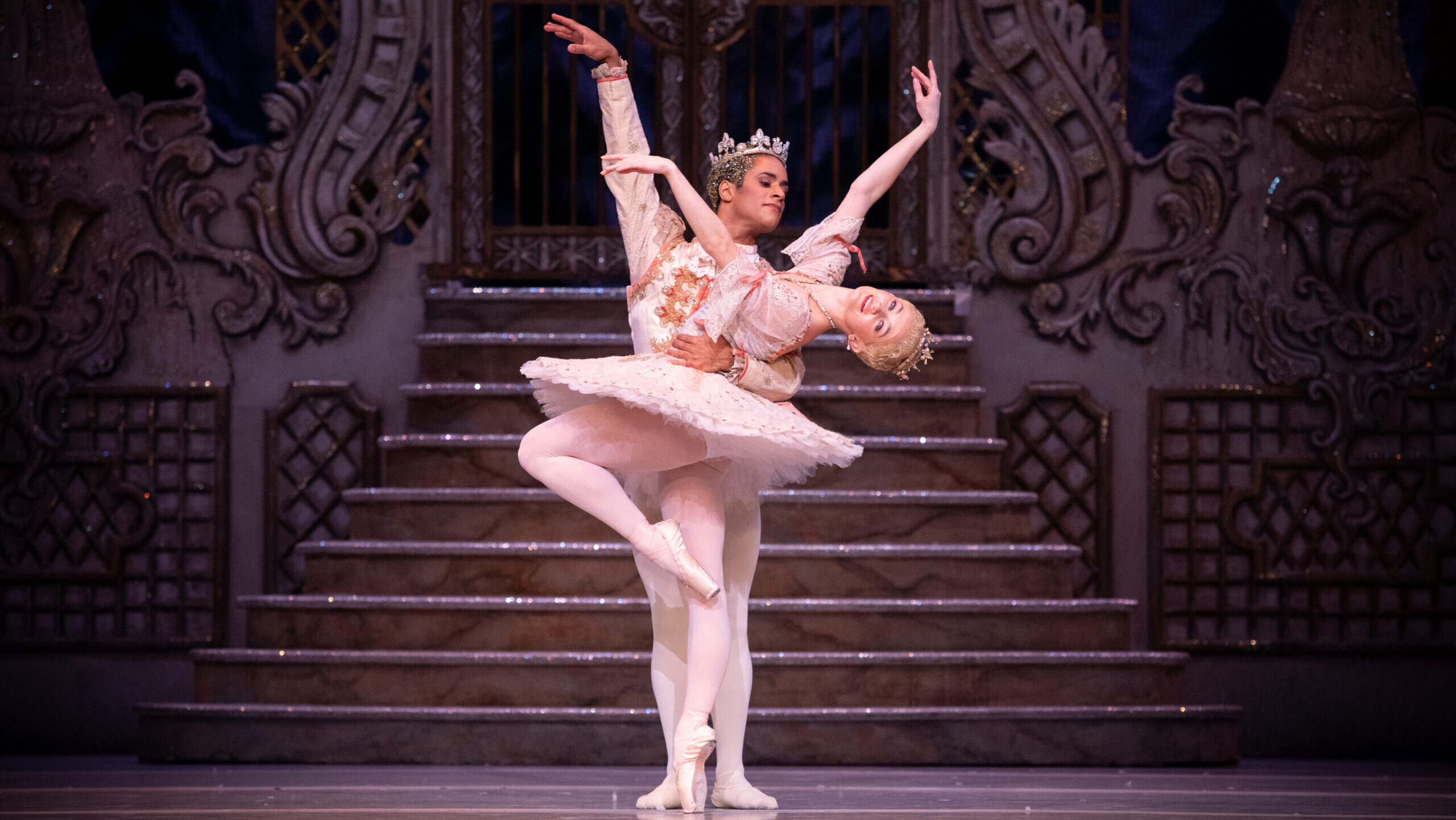 Anna Rose O'Sullivan poses in parralel passee on pointe, bending her upper body back in cambé with her arms in third arabesque. Marcelino Sambé stands beihnd her and holds her by the waist. She wears a pink tutu and gold tiara, while he wears a pink tunic, white tights and slippers, and a large crown.