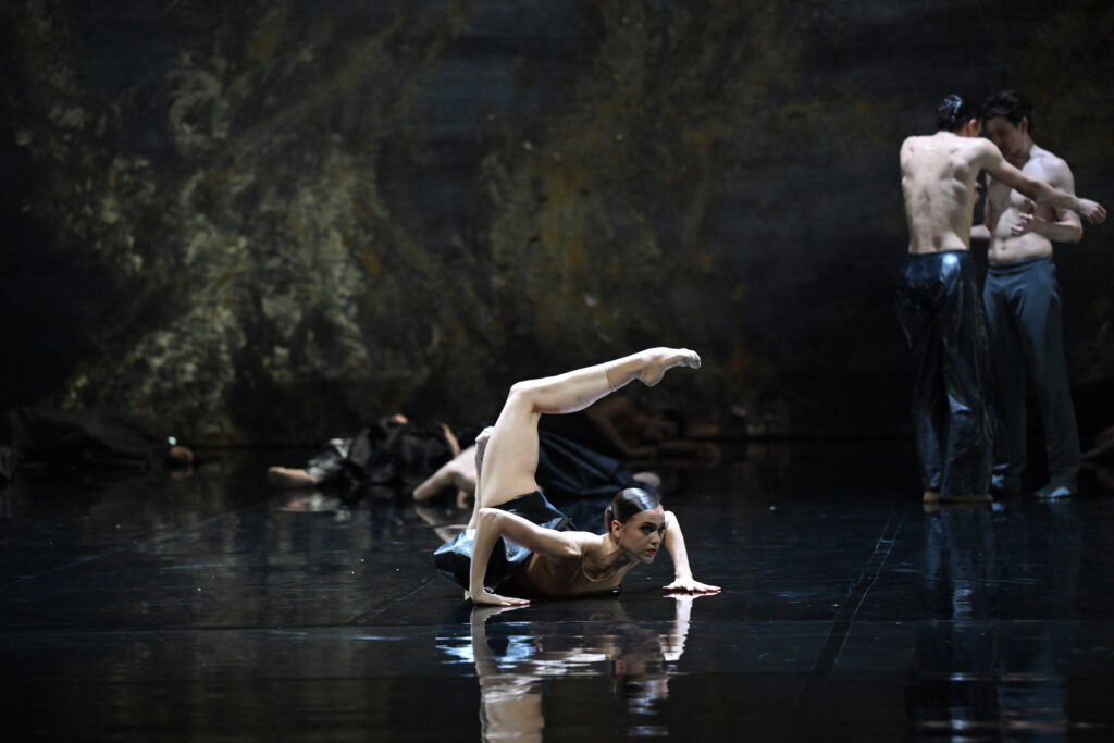 Mackenzie Brown lays on her stomach onstage with her arms out by her sides, elbows bent and hands flat on the floor. She wears a gold leotard and black skirt and lifts her right leg behind her in an attitude position.