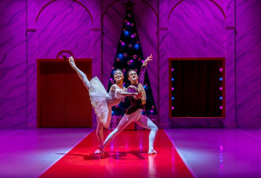 Ryoko Yagyu does a penché arabesque on pointe while holding on to the right shoulder and hand of her partner, who lunges to his left. She wears a pink tutu and tiara, while her partner wearsa a dark purple tunic, white tights and white slippers. They perform in front of a Christmas tree in a large marble foyer.