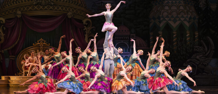 A large group of female ballet dancers dressed as flowers surround a cavalier lifting the Dew Drop Fairy, who poses in a parallel retiré.