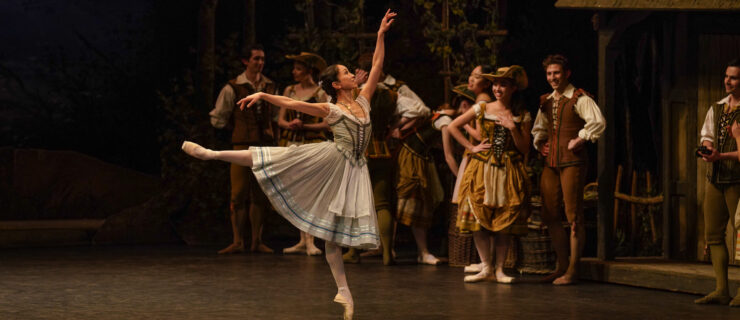 Dressed as Giselle in a light blue and white peasant dress, Erina Takahashi stands on pointe in arabesque. She smiles as she looks up to her front hand as a crowd of corps members watch and smile.