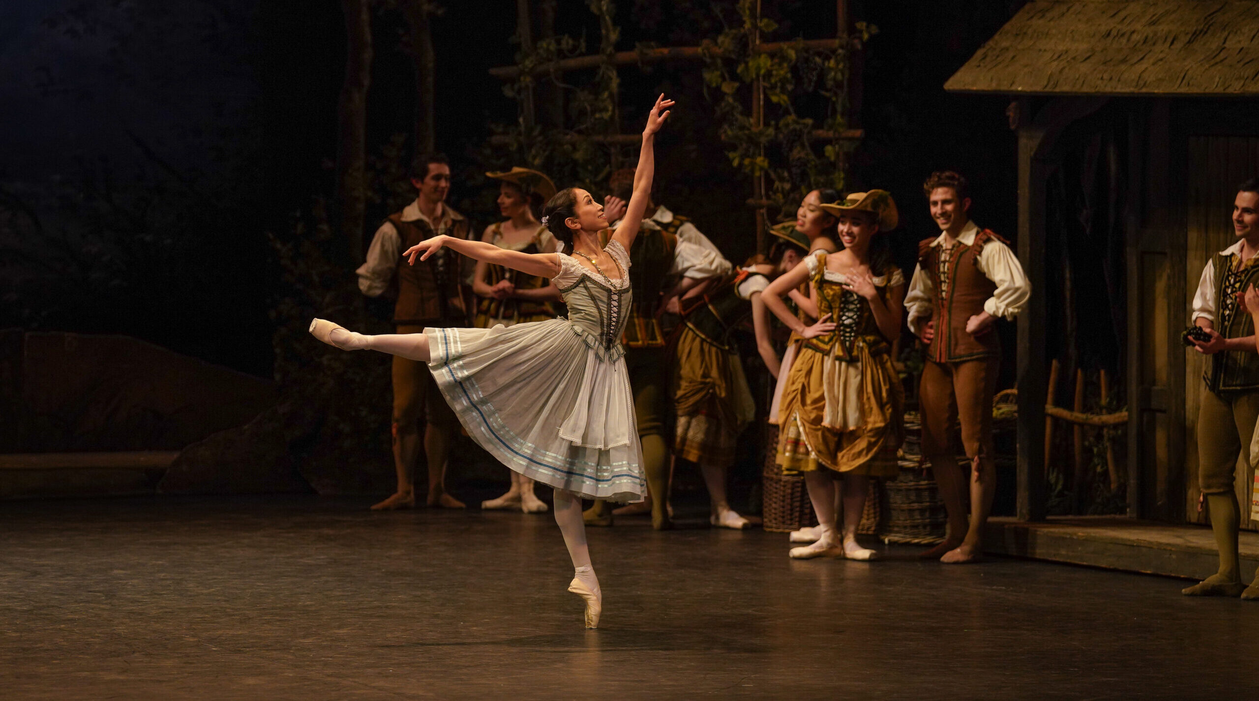 Dressed as Giselle in a light blue and white peasant dress, Erina Takahashi stands on pointe in arabesque. She smiles as she looks up to her front hand as a crowd of corps members watch and smile.