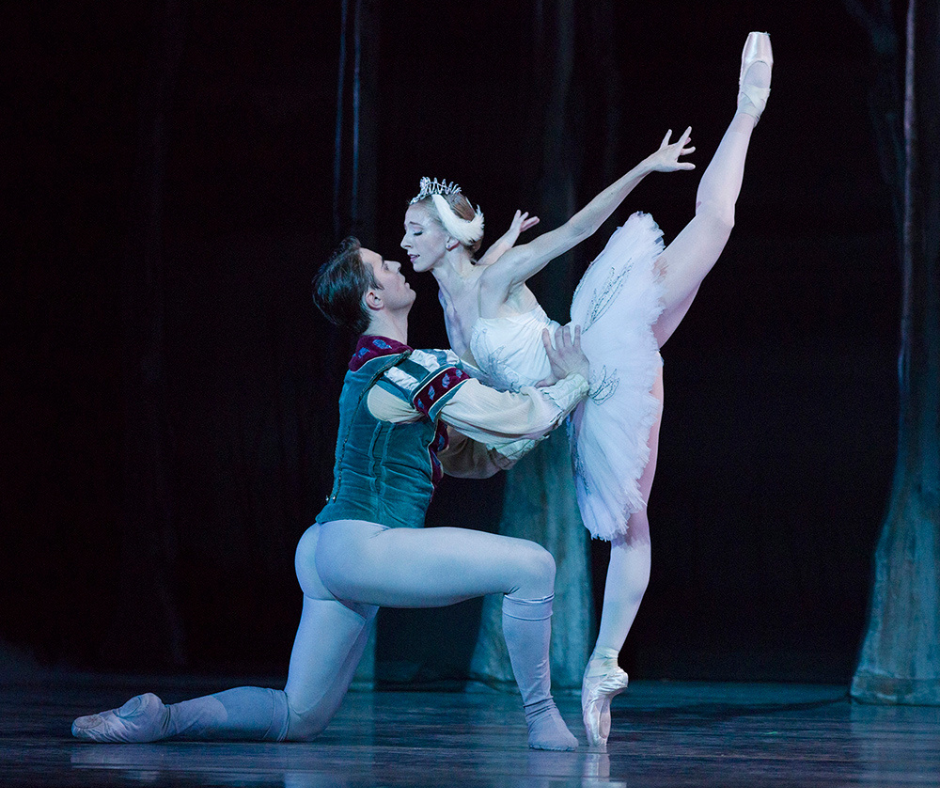 Jessica Lind, as Odette, stands on pointe in a penché attitude facing her partner, Luke Ingham, who kneels and supports her around the waist. He wears a teal blouse and light gray tights, and she wears a white pancake tutu and swan crown.