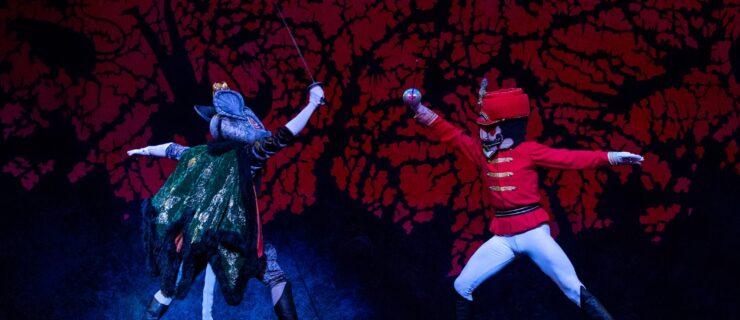 Two male dancers, one is a rat costume and the other dressed as a Nutcracker, lunge towards each other with swords in front of a black and red backdrop.