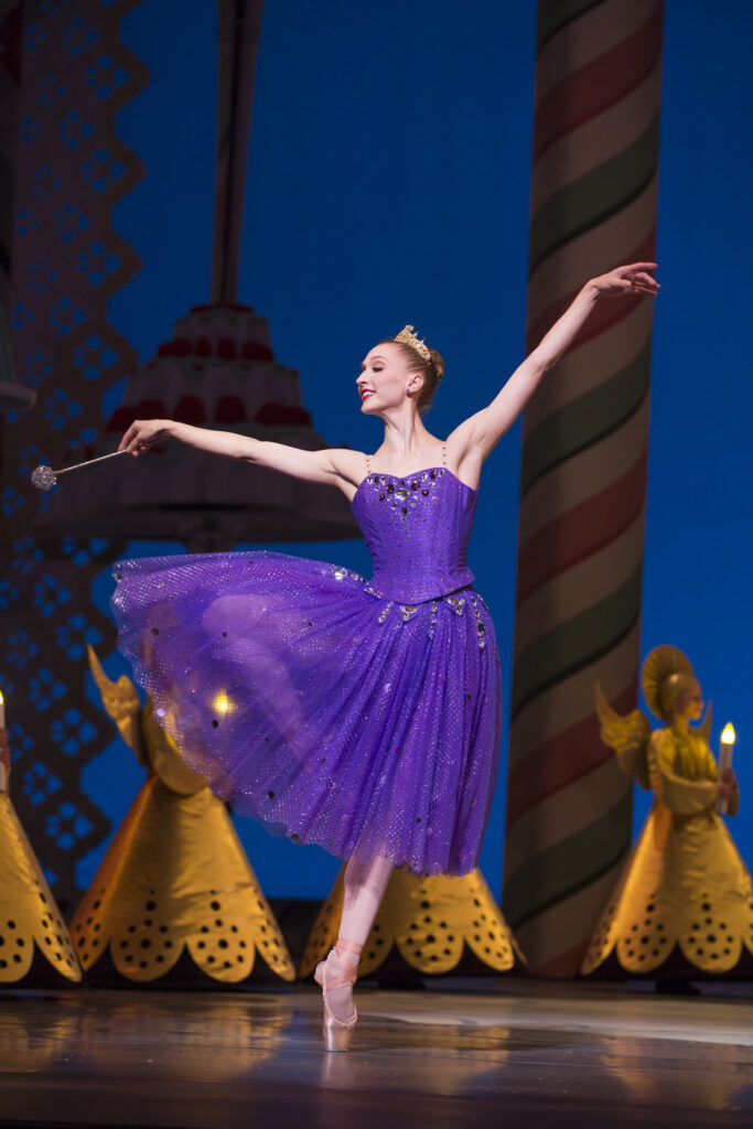 Elizabeth Murphy, wearing a dark purple, knee-length tutu and pink pointe shoes, does a relevé passé with her right leg and opens her arms out, holding a silver wand in her right hand.