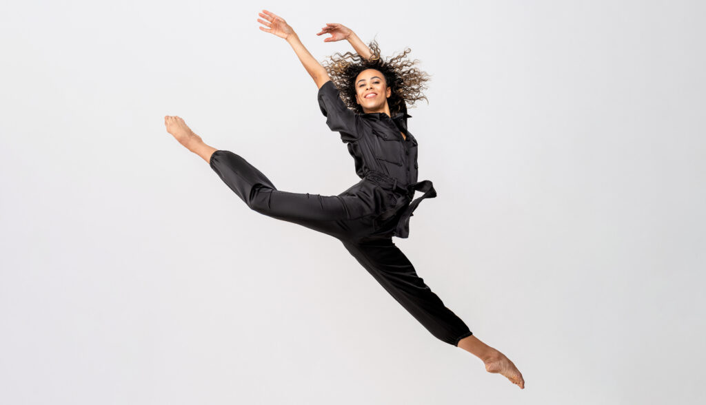 Nikisha Fogo poses in a split leap with her back leg in attitude, her arms raised overhead and flipped to the back. She grins at the camera, her long dark hair flying. She wears a dark gray belted jumpsuit.