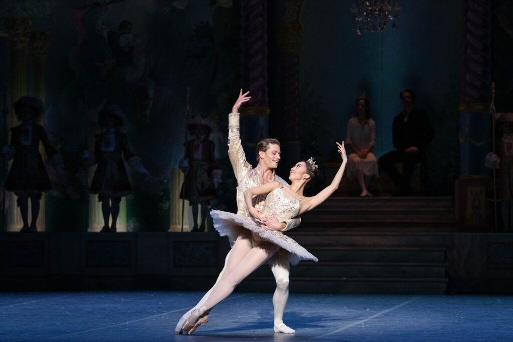 Chisako Oga and Derek Dunn dance a pas de deux onstage. Dunn, in a white tunic and tights, holds Oga by the waist as she poses in sus-sous and dips her slightly to his left. She wears a white tutu and silver tiara and looks up at him, raising her left amr to match his raised right arm.