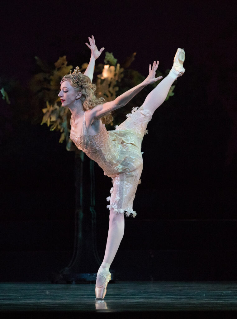 Jessica Lind penchés in arabesque on pointe with her arms extended behind energetically. Her long blond hair is curled and down, and she is dressed as a fairy queen in a short gold and cream dress and a gold crown.