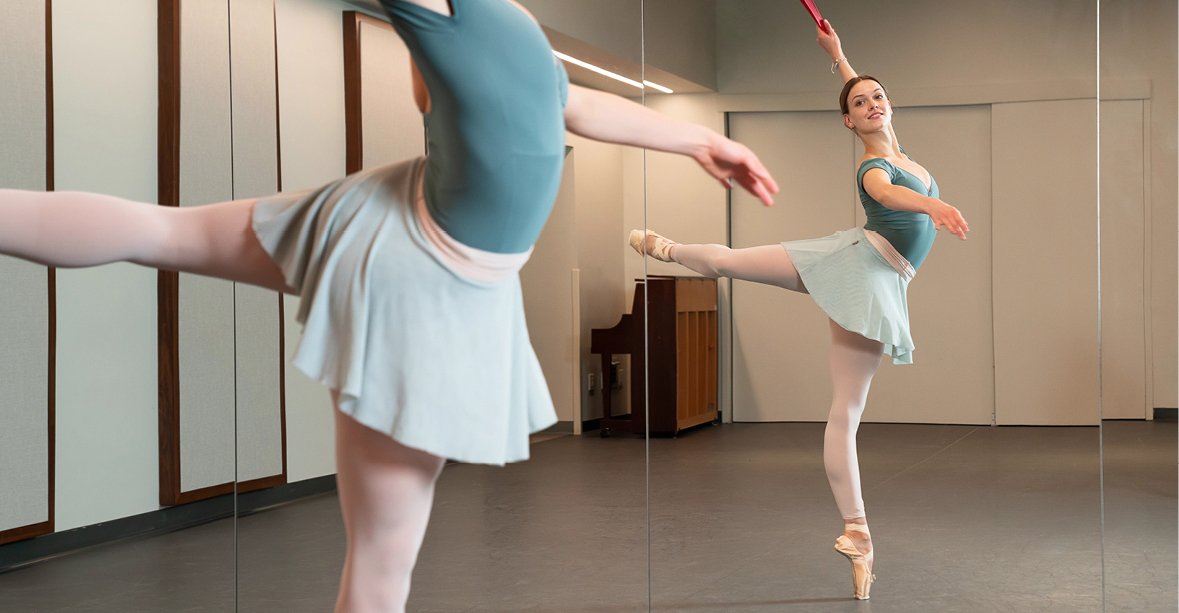 Mackenzie Brown does an attitude on pointe in a large dance studio. The photo shows a close of of her torso as well as her full body's reflection in the mirror. She wears a green leotard, light green skirt, and pink tights.