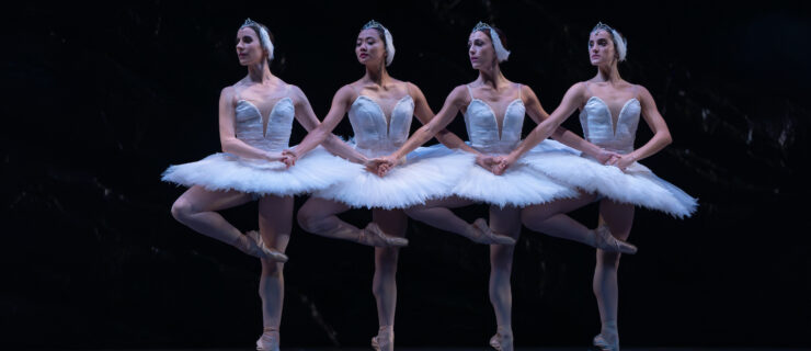 Four ballerinas in identical white tutus and feathered headpieces join hands and do a relevé passé with their right leg. They all look over their right shoulder and perform in front of a dark backdrop.