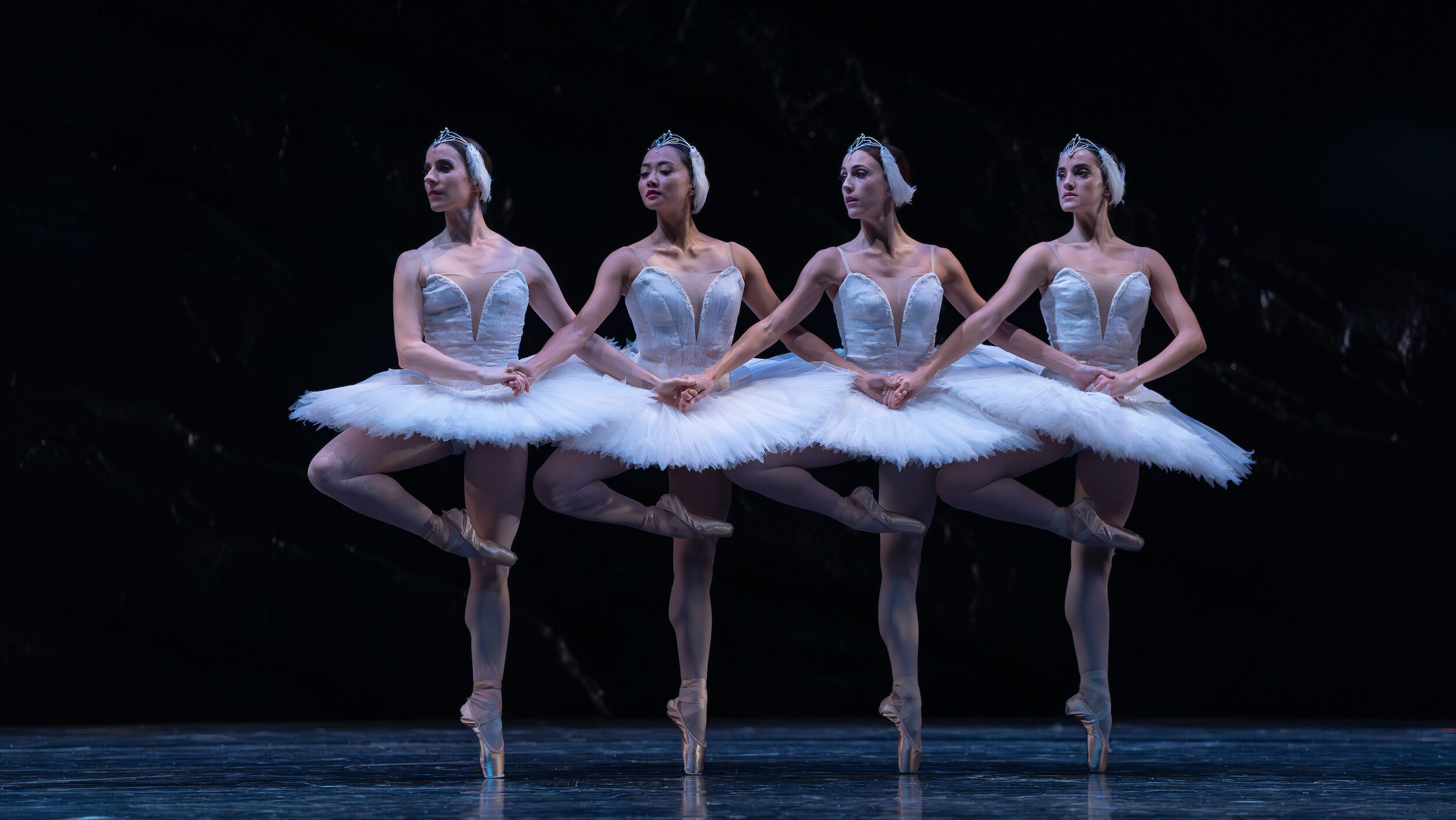 Four ballerinas in identical white tutus and feathered headpieces join hands and do a relevé passé with their right leg. They all look over their right shoulder and perform in front of a dark backdrop.