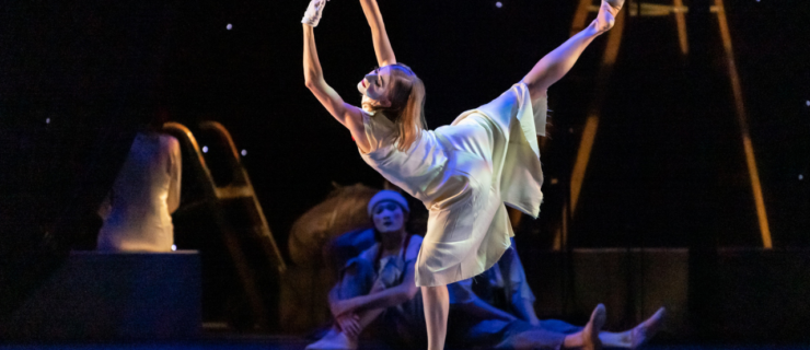 Jessica Lind wears a flowing white dress, ballet shoes, and white gloves as she dances onstage. She arabesques with her supporting leg in plié, arms in a high fifth with her fingers touching. Her blonde hair is down, and she wears white makeup on her face for a mime-ish or clownish effect.