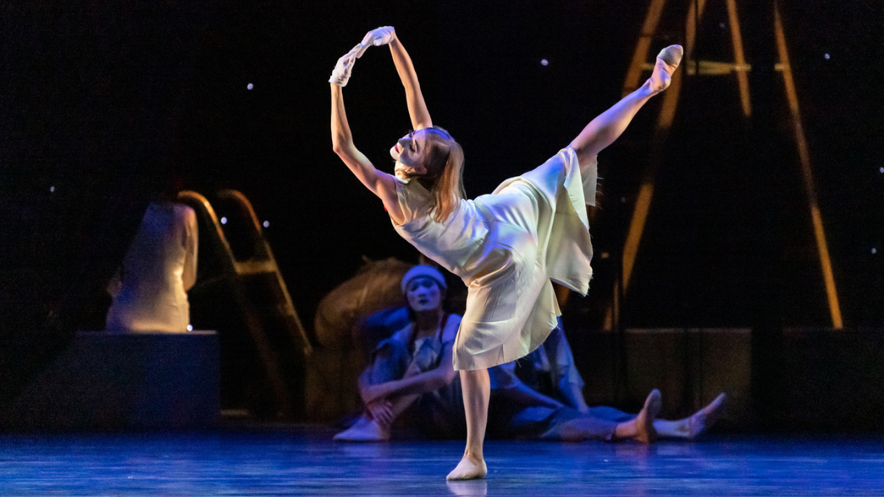 Jessica Lind wears a flowing white dress, ballet shoes, and white gloves as she dances onstage. She arabesques with her supporting leg in plié, arms in a high fifth with her fingers touching. Her blonde hair is down, and she wears white makeup on her face for a mime-ish or clownish effect.