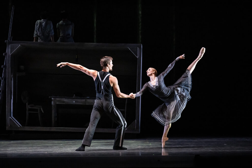 Jessica Lind and John-Paul Simoens dance together on a dark stage in dark gray and black costumes. He stands in fourth and holds her hand as she faces him and penchés on pointe in arabesque.