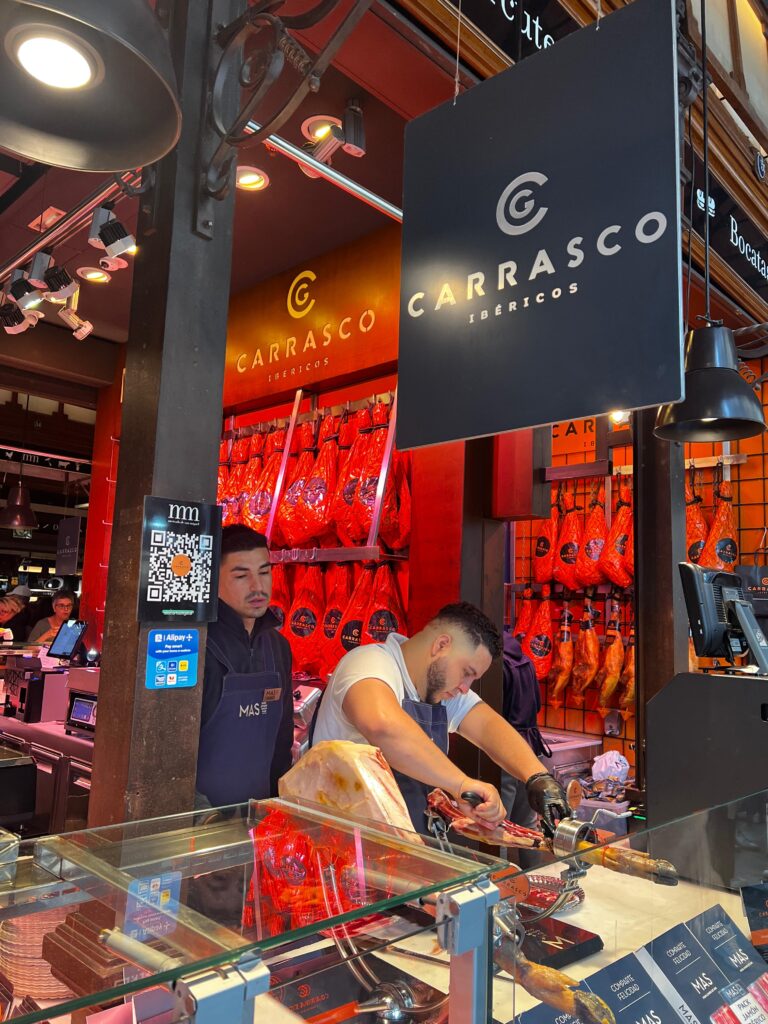 A young man slices jamón at a meat counter while another young man stands behind him and watches.