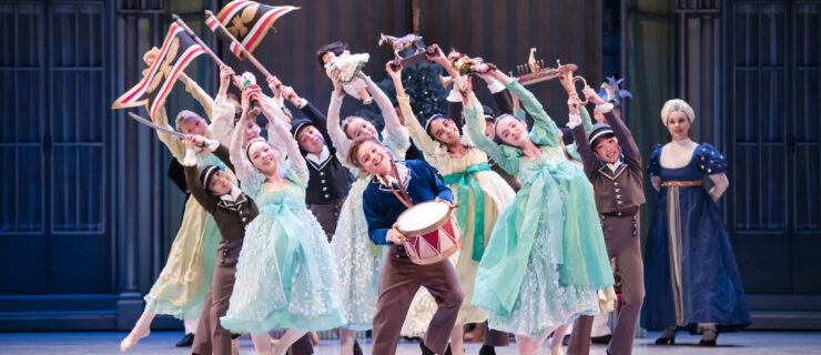 During a performance of the Nutcracker, group of about a dozen children in 19th century costumes face front and dance close together, leaning their bodies to their right with their arms up. Some carry dolls and toys while others carry flags, while a boy in the front holds a red and white drum. They all smile brightly.