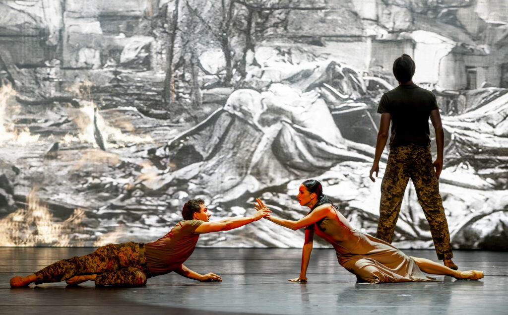 In front of a projection of war-torn Ukraine, three dancers perform onstage. In the front, a male and female dancer lie on their sides and reach toward each other, the man holding the woman's wrist as she looks at him in alarm. In the back, a second man stands with his back to the audience, looking at the projection in an active stance. The men wear camo-print pants and olive-colored t-shirts, and the woman wears a long tan sleeveless dress.