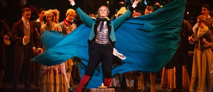 A male dancer wearing a gray wig, short turquoise coat, blue knickers, red socks and a blue cape, stands center stage and throws his arms up to present a gift on the fllor. He smiles towards the audience and his cape flies wide behind him. Other dancers in period costumes milll about behind him, watching.