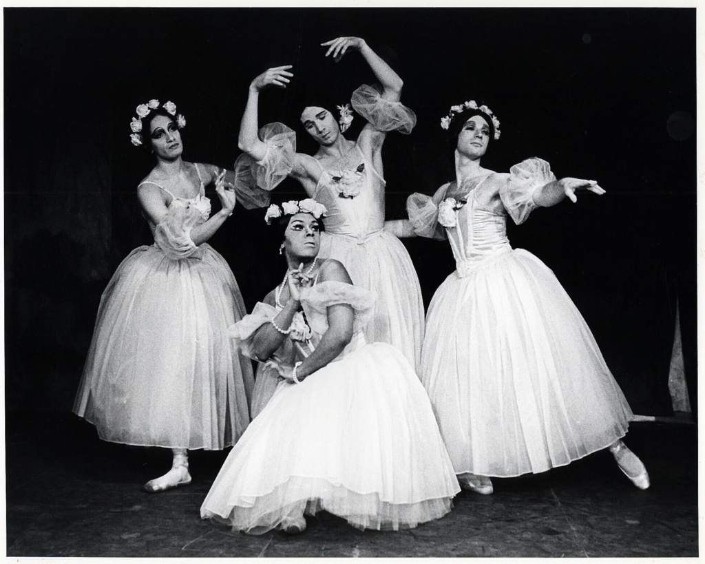 A black and white archival image showing four dancers dressed in drag for a performance of "Pas de Quatre." They wear long romantic tutus and flower crowns, posing together in a tableau.