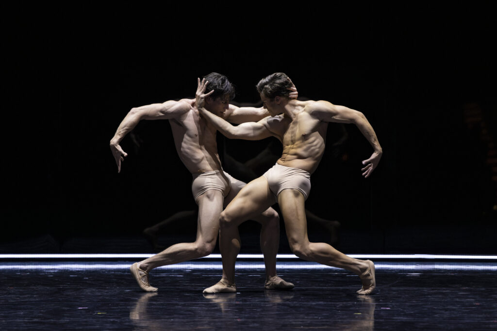 Onstage in stark lighting, two men mirror each other in an extreme undercurve position, legs staggered in parallel in plié, with one arm curved down behind them and the other grasping their partner's face. They wear nude-colored shorts.