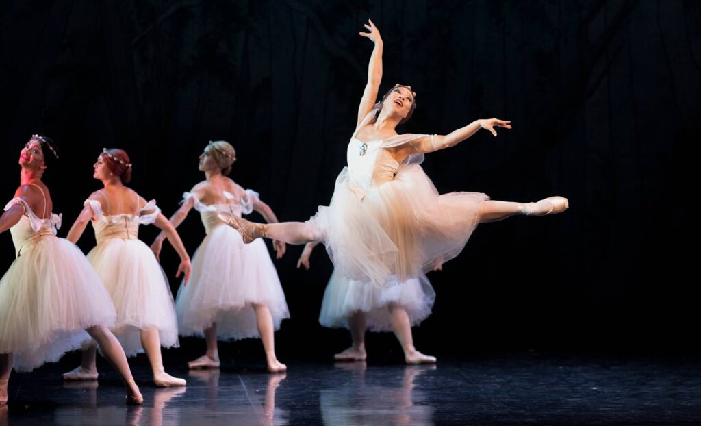 Onstage, dancers in drag perform "Les Sylphides" in long white romantic tutus. In the front, one dancer flies in a majestic saut de chat. Behind, four dancers stand in a lunged fourth position, their backs to her on the diagonal.