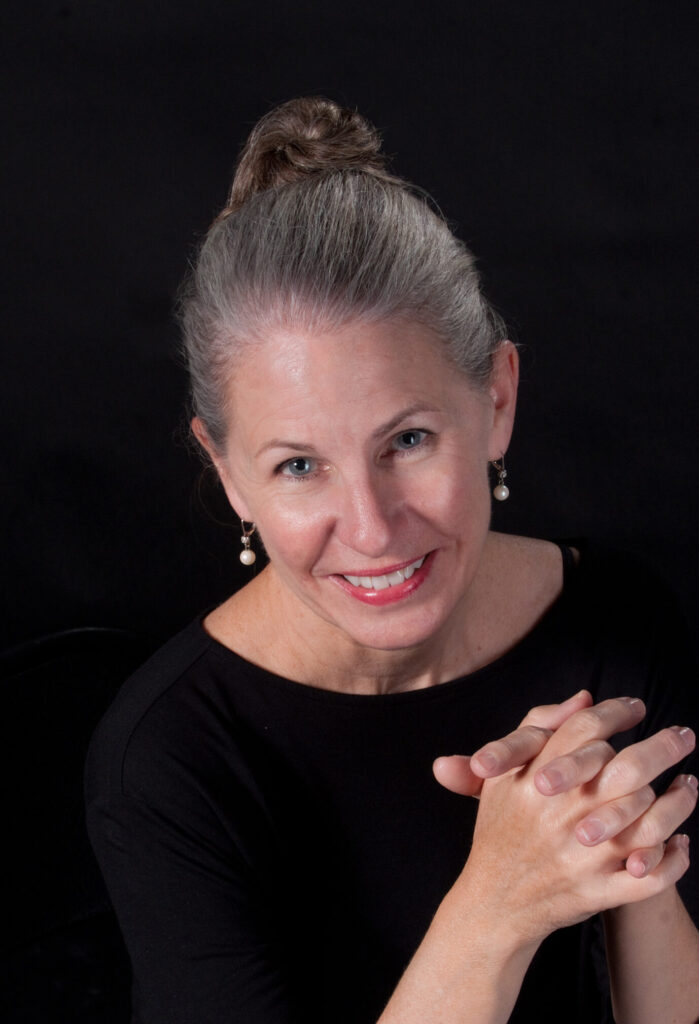 Colleen Smith, photographed from the chest up, wears a black sweater and pearl earrings and her hair in a high bun. She clasps her hands and smiles directly towards the camera.