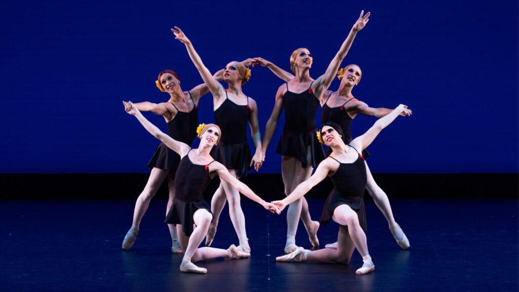 A group of six dancers in drag pose onstage in a tableau, four on the outside holding hands around two in the middle. They wear short black dresses with yellow flowers in their hair.