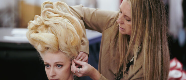 A woman with long brown hair stands over a dancer who is sitting down and pins a very tall blonde wig on her head.