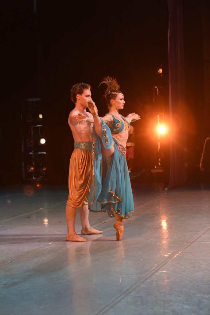 A male and female dancer perform a pas de deux onstage and are photographed from the wings. The woman, wearing a shirt blue top with gold detail, a long blue skirt, and feather headpiece, does a développé à la seconde on pointe with her right leg and her arms out to the side. Her partner, who wears knee-length gold pants with a turquise cummerbund, stands behind her and holds both of her hands.