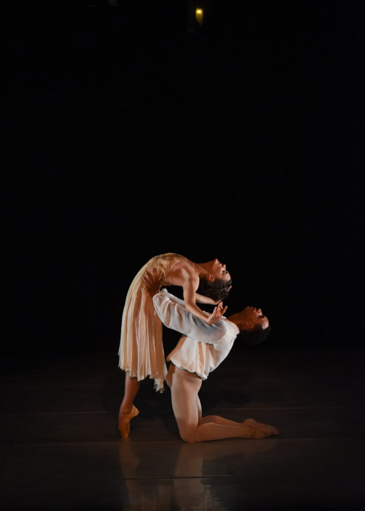 Lily Loveland, wearing a pink nightgown, stands in profile on pointe, her feet in parallel, and does a deep backbend. Her partner, Ihosvany Rodriguez, wearing a peasant blouse and tan tights, kneels behind her and holds her by the waist as he arches back.