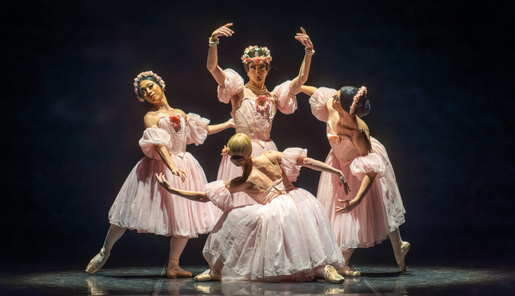 Four dancers dressed in drag perform "Pas de Quatre" in long pink romantic tutus and flower crowns. They pose in the piece's opening tableau.