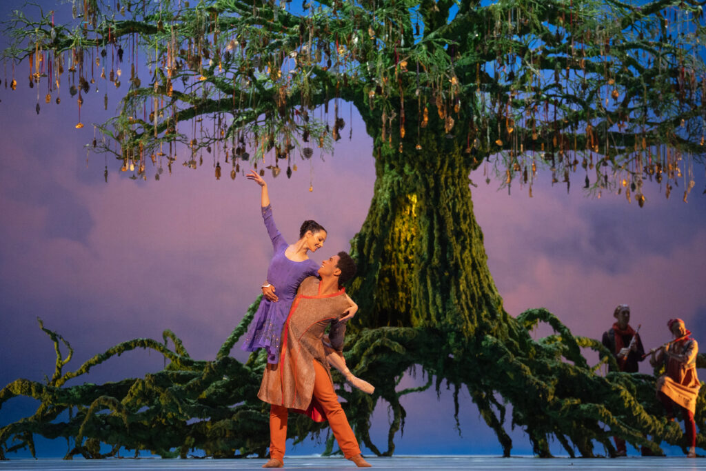 Marcelino Sambé, dressed in orange, lifts Francesca Hayward, dressed in a purple frock, on his right hip as they dance in front of a large tree with massive roots. Gold ornaments hang from the tree's branches above them.