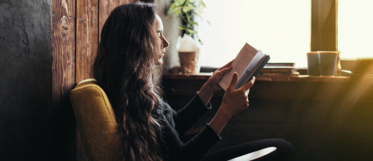 A young woman with long curly hair sitting in a comfortable armchair, reading a book.