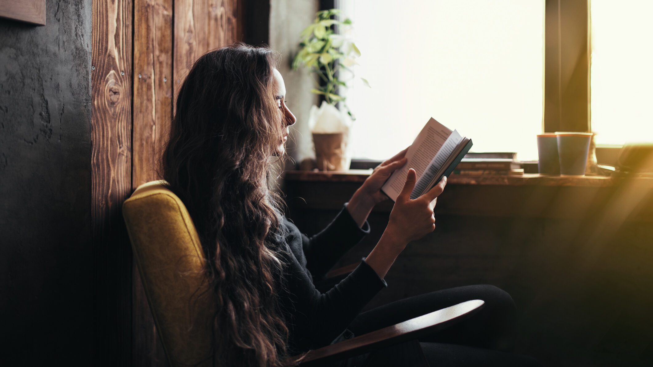 A young woman with long curly hair sitting in a comfortable armchair, reading a book.