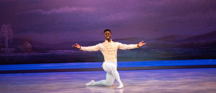 Brooklyn Mack kneels center stage during a performance and extends his arms out towards the audience, smiling widely. He wears a white tunic with gold trim, white tights, and white ballet slippers, and poses in front of a backdrop showing a lake and purple sky.