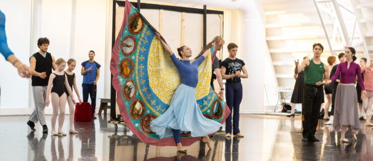 In a large ballet studio, a female dancer in a long sky-blue skirt stands confidently, arms lifted in a wide V. A large semi-circular cape is attached to her arms, which fans out behind her. Other dancers stand and watch behind her.