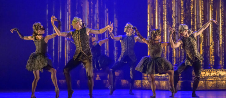 Three couples pose onstage, with either dancer facing the other. They plié and prop their right legs on forced arch, arms bent at the elbow as they gently overlap forearms. They wear masquerade-esque costumes with black and gold masks and silver and white wigs.