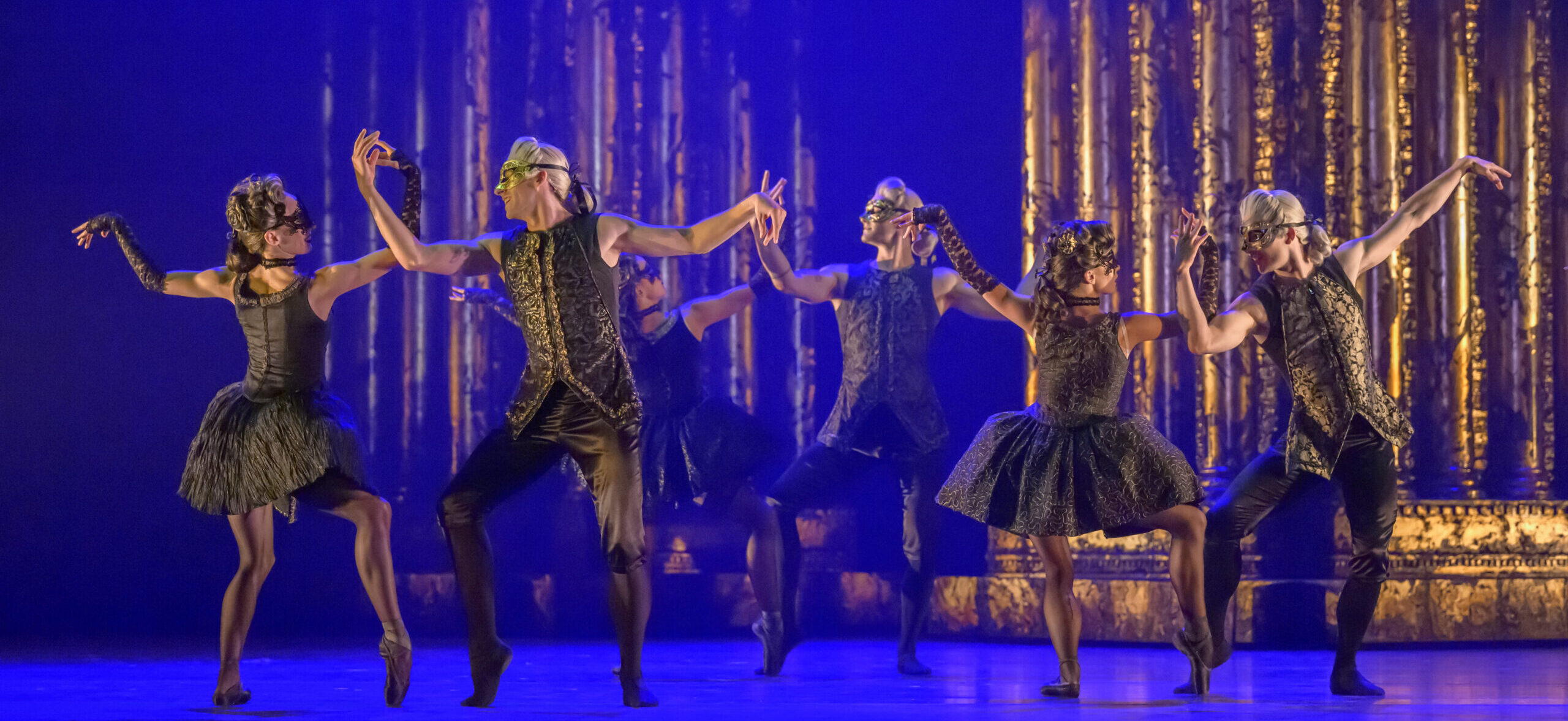 Three couples pose onstage, with either dancer facing the other. They plié and prop their right legs on forced arch, arms bent at the elbow as they gently overlap forearms. They wear masquerade-esque costumes with black and gold masks and silver and white wigs.