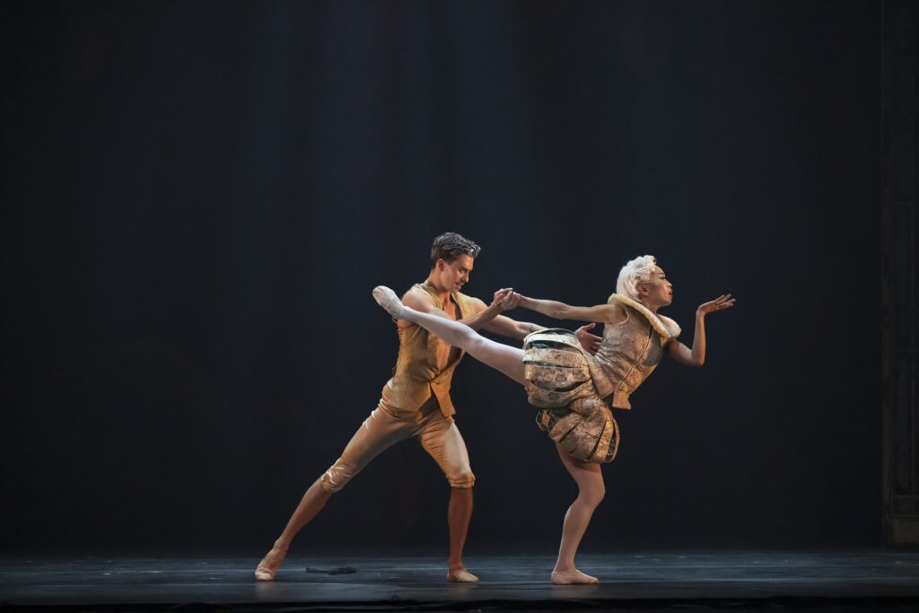 Dancing a pas de deux together, a ballerina pliés on her supporting leg as she reaches back for her partner's hand in arabesque. Her front arm bends as if to blow a kiss. Her partner lunges in toward her, supporting her waist with his other hand. They wear period-piece costumes in bright gold.