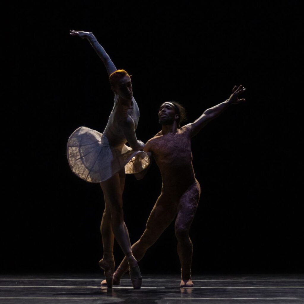 Two dancers perform onstage together in dark lighting. Jillian Davis stands on pointe about to step onto her right leg, torso twisted with her left arm extended up and back, and her right bend inward to hold Joe Gonzalez' hand. Gonzalez leans in toward Davis in a parallel side lunge, left arm extended outward.