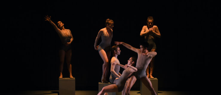 A group of six dancers in taupe leotards perform onstage in dim lighting. Three women stand on tan square boxes, while a male dancer sits on the edge of the box in the middle. In front of them a male and female dancer perform a duet.