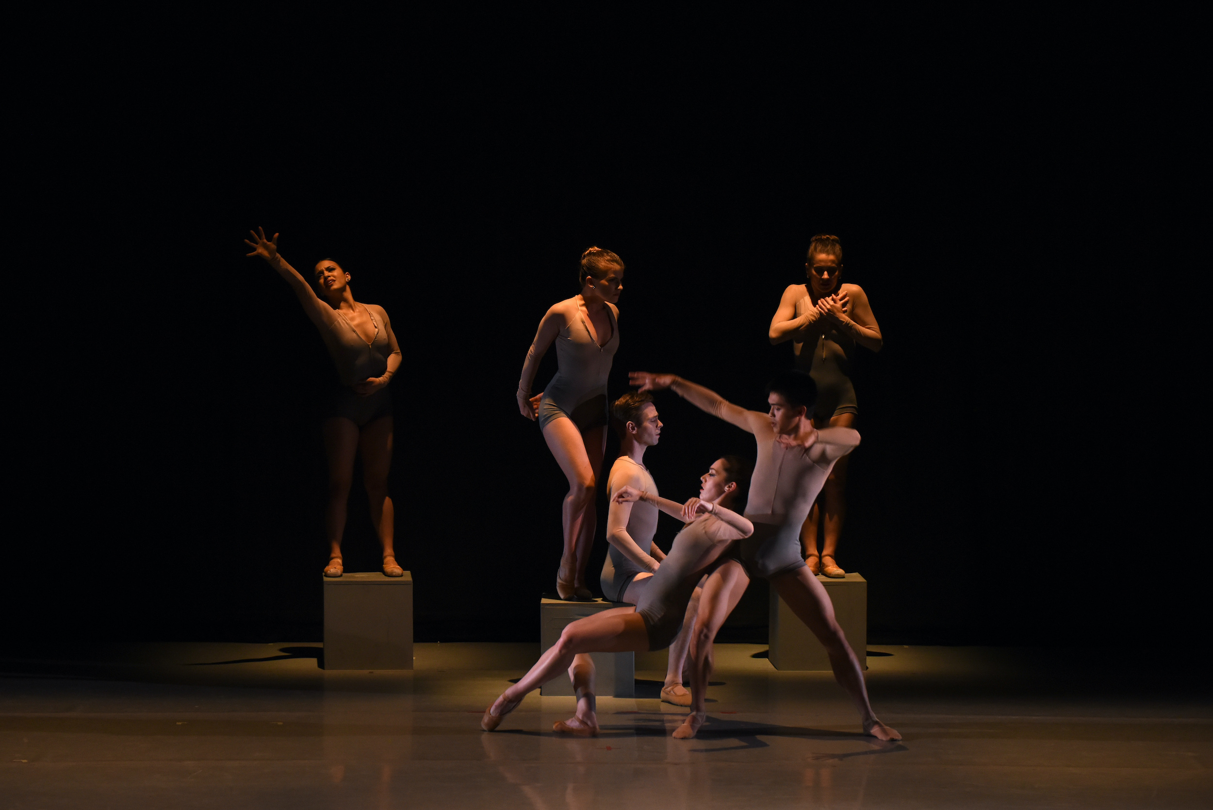 A group of six dancers in taupe leotards perform onstage in dim lighting. Three women stand on tan square boxes, while a male dancer sits on the edge of the box in the middle. In front of them a male and female dancer perform a duet.