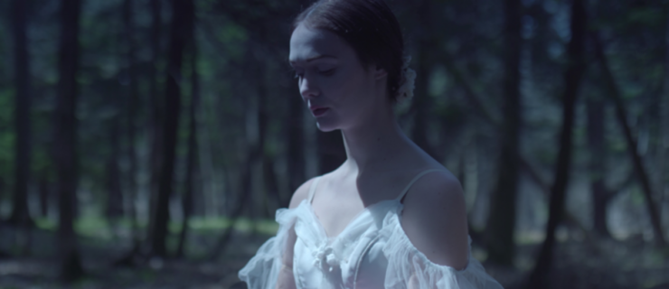 A close-up shot of Jurgita Dronina dressed as Giselle in a white dress with ruffled off-the-shoulder accents and a low bun. She crosses her arms in front, fingers turned up in the classic "willi" position. She looks down pensively.