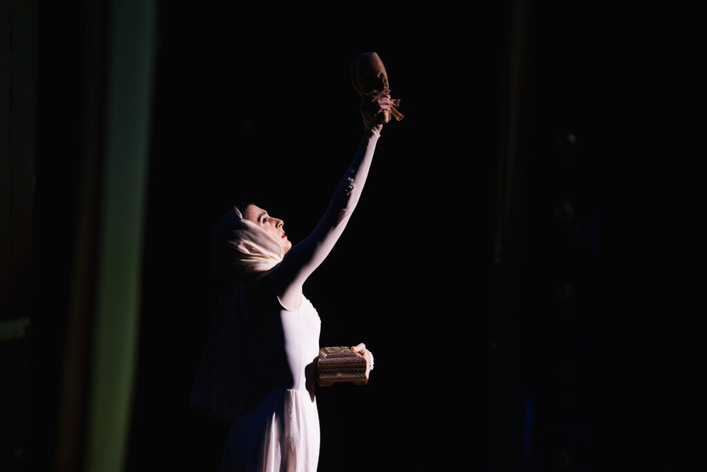 Tara Ghassemieh, shown from the hips up, wears a long white dress and veil, holding a wooden box close in her left hand and holding up a pair of flesh-colored pointe shoes in her right. She looks up to them, lit from the front.