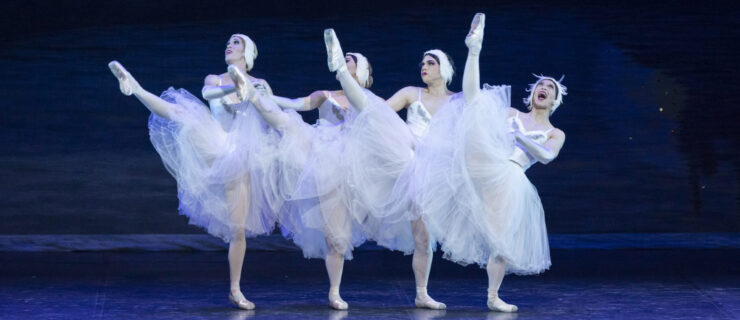 Four dancers dressed in drag as the four little swans from "Swan Lake" battement devant flamboyantly as they hold hands. They wear long white romantic tutus, pink tights, pointe shoes, and white feather headpieces.