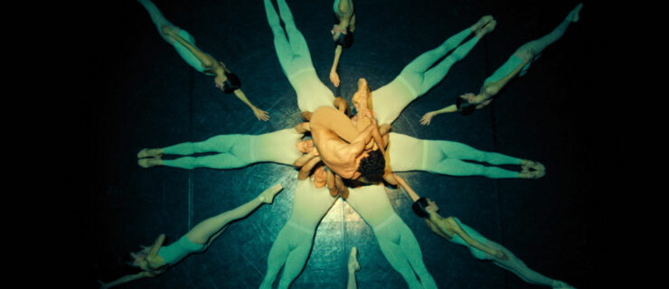 With the camera above, facing the floor, six dancers in white unitards lie on their backs in a circle, heads together, as they bend their arms up to support one man who is curled into a ball in the middle. He wears nude-colored tights. In the blank spaces of the six dancers, six other dancers in pointe shoes, white leotards, and tights lie on their sides with one arm raised to made additional spokes.